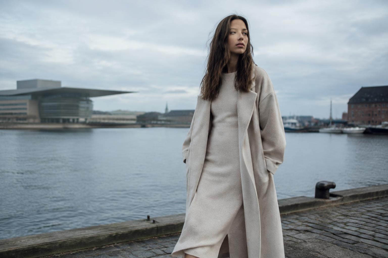 Woman in an oatmeal knit dress and long coat on a Copenhagen harbourfront